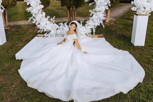 ein Brünette Braut im ein Schleier und ein Tiara im ihr Haar sitzt auf ein Verbreitung Kleid, posieren in der Nähe von ein Weiß Bogen gemacht von Blumen im das gestalten von ein Kreis. auf ein Weiß Hintergrund. sonnig Tag. Hochzeit Zeremonie foto