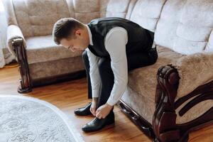 ein Mann ist Putten auf seine Schuhe während Sitzung auf ein Sofa im ein Hotel Zimmer. ein jung Unternehmer ist vorbereiten zu gehen auf ein Geschäft Reise. Geschäftsmann vorbereiten zum Reise im Zuhause Schlafzimmer früh im das Morgen foto