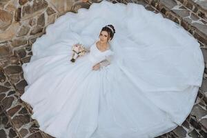 ein Brünette Braut mit ein Tiara im ihr Haar ist Lügen auf ein Kleid gelegt aus im das gestalten von ein Kreis, posieren während halten ein Strauß. auf ein Weiß Hintergrund. sonnig Tag. Hochzeit Zeremonie foto