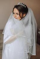 Porträt von ein unglaublich schön Mädchen Braut im ein Weiß Kleid im das Schlafzimmer, das Braut posiert halten ihr Schleier im ihr Hände und Abdeckungen Sie selber mit Es. foto