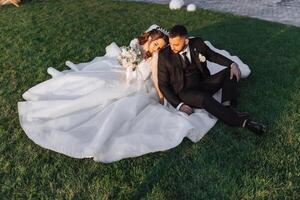 Porträt von schön Hochzeit Paar Sitzung auf Grün Gras im Park im Sommer. jung heiter Frau Braut umarmen Bräutigam. foto