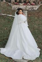 ein Brünette Braut im ein Weiß Kleid mit ein lange Zug hält das Kleid und Spaziergänge Nieder das Pfad bedeckt mit Herbst Blätter. Hochzeit Foto Session im Natur. schön Haar und bilden. Feier