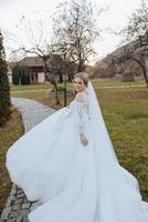 ein blond Braut im ein Weiß Kleid mit ein lange Zug hält das Kleid und Spaziergänge entlang das Stein Weg. Herbst. Hochzeit Foto Session im Natur. schön Haar und bilden. Feier