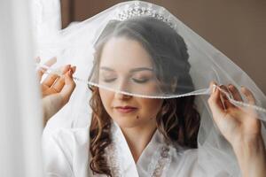 Porträt von ein unglaublich schön Mädchen Braut im ein Weiß Kleid im das Schlafzimmer, das Braut posiert halten ihr Schleier im ihr Hände und Abdeckungen Sie selber mit Es. foto