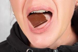 ein Frau, deren Gesicht ist nicht sichtbar, hält ein Stück von Schokolade im ihr Mund. foto