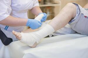 Krankenschwester Bandagen das Bein. Fraktur von Mensch niedriger Glieder. Behandlung von gebrochen Knochen. auferlegen ein Gips. geduldig chirurgisch Abteilung. das Arzt Hände spannen das Binde auf seine Bein foto