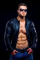 muskulös und passen jung Bodybuilder. Fitness männlich Modell- posieren Über dunkel Hintergrund. Leder Jacke auf nackt Torso. foto