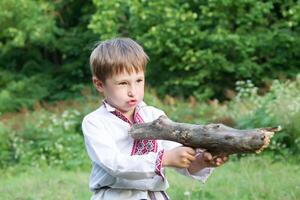 slawisch Kind im National Kleidung. Belarussisch Junge Theaterstücke mit ein Stock wie mit ein Pistole. selbstgemacht hölzern Spielzeuge. Kinder- Vorstellung. Russisch Mann. foto