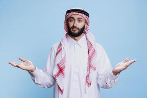 nachdenklich Mann tragen Muslim thobe und Kopftuch, Stehen und Achselzucken Schultern während suchen beim Kamera. arabisch Person zeigen Zögern mit öffnen Waffen Geste Studio Porträt foto