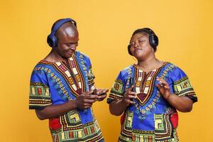 afrikanisch amerikanisch Paar genießen Musik- zusammen mit Kopfhörer und Smartphone. Frau Hören zu Wiedergabeliste mit geschlossen Augen und zufrieden Ausdruck während Mann drehen auf Lied auf Handy, Mobiltelefon Telefon foto