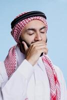 schläfrig Mann mit geschlossen Augen tragen traditionell Muslim Kopfschmuck und thobe Gähnen. erschöpft arabisch gekleidet im islamisch Kleider Abdeckung Mund mit Arm während Gefühl Schläfrigkeit und Müdigkeit foto