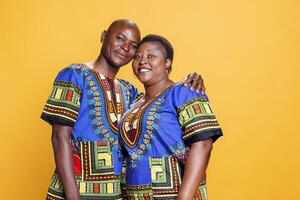 lächelnd schwarz Mann und Frau Paar gekleidet im ethnisch Kleider umarmen, zeigen Liebe und Glück Studio Porträt. Ehefrau und Mann und suchen beim Kamera mit heiter Ausdruck foto