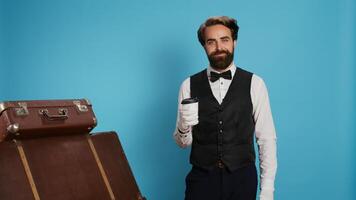 Hotel Porter genießen Kaffee Tasse auf Kamera, Page tragen Fachmann nobel Uniform und Handschuhe. stilvoll Türsteher Trinken Erfrischung, posieren mit Gepäck auf Blau Hintergrund. foto