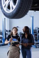 Mechaniker beim Auto Reparatur Geschäft führt jährlich Fahrzeug Untersuchung, informieren Klient Über erforderlich Räder Ersatz. Garage Arbeiter Gespräche mit Kunde unterhalb Auto nach Fertigstellung Inspektion foto