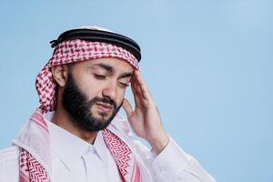 Muslim Mann im traditionell Kleider ausdrücken Beschwerden und Reiben Tempel während Leiden von Migräne Attacke. arabisch Person tragen ghutra Kopfschmuck haben Kopfschmerzen und Gefühl Schmerzen foto