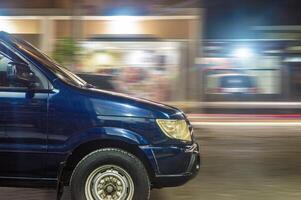 Schwenken Fotografie von ein dunkel Blau Kleinbus Fahren schnell beim Nacht im ein städtisch Bereich mit Geschäfte foto
