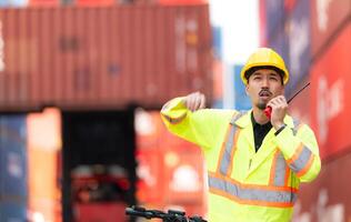Porträt von ein Warenhaus Arbeiter Sprechen auf ein Walkie Talkie mit ein Mitarbeiter im ein leeren Container Warenhaus während Fahren ein Roller zu prüfen Arbeit foto