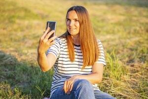 schön jung Frau mit Handy, Mobiltelefon Telefon Sitzung auf Grün Gras draußen foto