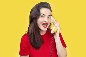 ein schön Brünette Frau mit rot Lippenstift spricht auf ein alt Gelb Telefon mit ein Kabel. sie ist lächelnd. auf ein Gelb Hintergrund foto