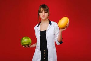 ein weiblich Arzt Krankenschwester im ein Weiß Mantel mit Obst im ihr Hände posiert auf ein rot Hintergrund, Melone, Wassermelone, foto