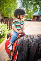 schön wenig Mädchen zwei Jahre alt Reiten Pony Pferd im groß Sicherheit Jockey Helm posieren draußen auf Landschaft foto