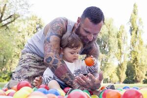 tätowiert Vater haben Spaß mit seine Sohn im das Park mit farbig Bälle foto