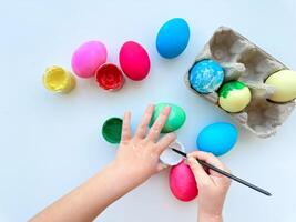 Kinder Hände Farbe Ostern Eier mit Bürste umgeben durch bunt Eier und Gläser von Farben auf Weiß Tisch, kreativ Urlaub Aktivität. foto