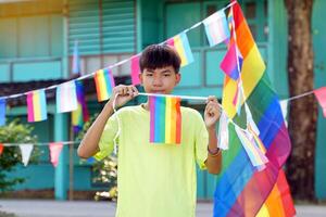asiatisch Junge hält ein Regenbogen Flagge im Vorderseite von ein Haus dekoriert mit Regenbogen Flaggen während Stolz Monat zu Show lgbt Stolz und Identität. Sanft und selektiv Fokus. foto