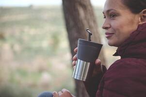 Nahansicht von ein glücklich lächelnd Frau Trinken heiß Tee oder Kaffee von ein Reise Becher während ruhen nach Wandern im Natur foto