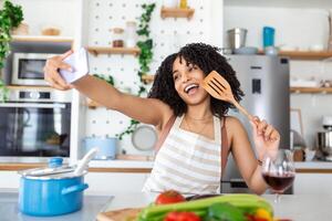 Porträt von lächelnd jung afrikanisch amerikanisch Frau nehmen Selfie mit Smartphone während Kochen im Küche beim Zuhause foto