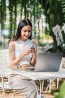 ein jung Frau ist absorbiert im SMS auf ihr Smartphone, mit ein Laptop und Kaffee beim ein draussen Tabelle umgeben durch Grün. foto