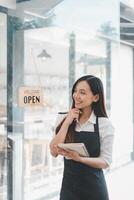 schön jung Barista Frau im Schürze halten bestellen Papier und Stehen im Vorderseite von das Tür von Cafe mit öffnen Zeichen Tafel. Geschäft Inhaber Anfang Konzept. foto