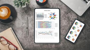 Büro Schreibtisch Arbeitsplatz mit Geschäft Diagramm auf Bildschirm Tablette und Smartphone, Laptop, Stift, Notizbuch, Brille und Tasse von Kaffee, oben Aussicht eben legen. foto