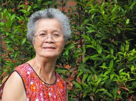 Senior asiatisch Frau mit kurz Weiß Haar lächelnd und suchen beim das Kamera während Stehen im ein Garten. Raum zum Text. Konzept von alt Menschen und Gesundheitswesen foto