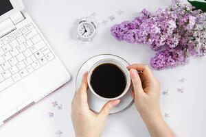 weiblich Hände halten Tasse von Espresso Kaffee. Alarm Uhr und lila Blumen auf Licht Hintergrund. foto