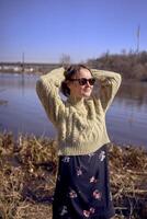 stilvoll jung Frau im ein Licht Grün Sweatshirt und Sonnenbrille auf das Bank von ein Frühling Fluss foto