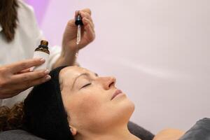 Ernte Kosmetikerin bewirbt sich Serum auf Klient Gesicht im Schönheit Salon foto