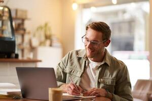 Porträt von modern Mann im Gläser, Sitzung im ziemlich leeren Cafe mit Laptop, Schreiben Nieder Anmerkungen im Notizbuch, studieren online, Fernbedienung E-Learning, Vorbeigehen Netz Kurs foto