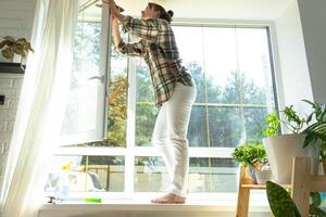 Frau manuell wäscht das Fenster von das Haus mit ein Lappen mit sprühen Reiniger und Mopp Innerhalb das Innere mit Weiß Vorhänge. Wiederherstellen bestellen und Sauberkeit im das Frühling, Reinigung warten foto