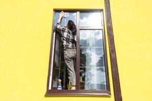 ein Frau manuell wäscht das Fenster von das Haus mit ein Lappen mit ein sprühen Reiniger und ein Mopp außen. Sicherheit beim Höhe, Wiederherstellen bestellen und Sauberkeit im das Frühling, Reinigung Bedienung foto