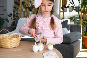 ein süß Mädchen mit Rosa Hase Ohren macht ein Ostern Kunst - - dekoriert ein Ei im das bilden von ein Einhorn mit Strasssteine, Horn, Blumen im das Innere von ein Haus mit Pflanzen. foto