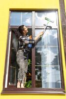 ein Frau manuell wäscht das Fenster von das Haus mit ein Lappen mit ein sprühen Reiniger und ein Mopp außen. Sicherheit beim Höhe, Wiederherstellen bestellen und Sauberkeit im das Frühling, Reinigung Bedienung foto