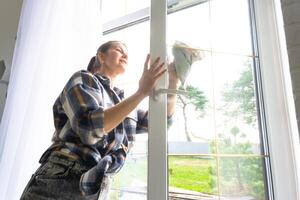 Frau manuell wäscht das Fenster von das Haus mit ein Lappen mit sprühen Reiniger und Mopp Innerhalb das Innere mit Weiß Vorhänge. Wiederherstellen bestellen und Sauberkeit im das Frühling, Reinigung warten foto