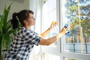 Frau manuell wäscht das Fenster von das Haus mit ein Lappen mit sprühen Reiniger und Mopp Innerhalb das Innere mit Weiß Vorhänge. Wiederherstellen bestellen und Sauberkeit im das Frühling, Reinigung warten foto