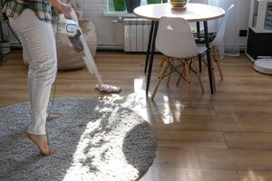 ein Frau Staubsauger ein runden Teppich im ein Haus unter Haus Pflanzen mit ein Hand Vakuum Reiniger. Allgemeines Reinigung von das Haus, Reinigung Bedienung und Hausfrau foto