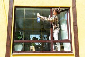 ein Frau manuell wäscht das Fenster von das Haus mit ein Lappen mit ein sprühen Reiniger und ein Mopp außen. Sicherheit beim Höhe, Wiederherstellen bestellen und Sauberkeit im das Frühling, Reinigung Bedienung foto