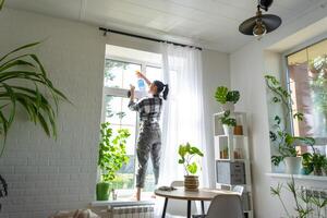 Frau manuell wäscht das Fenster von das Haus mit ein Lappen mit sprühen Reiniger und Mopp Innerhalb das Innere mit Weiß Vorhänge. Wiederherstellen bestellen und Sauberkeit im das Frühling, Reinigung warten foto