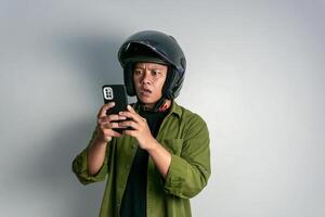 Erwachsene asiatisch Mann verwirrend wann suchen zu seine Handy während tragen Motorrad Helm. Mann suchen zu seine Telefon mit schockiert Ausdruck foto