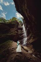 ein jung Mann und seine Ehefrau sind Stehen im ein Umarmung im ein Schlucht gegen das Hintergrund von ein hoch Wasserfall. Jungvermählten Hochzeit Paar im das Berge foto