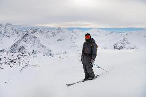 jung Mann steht auf ein Felsen beim das oben von ein Berg mit ein Rucksack und Ski, Aufpassen ein lange alpin Panorama foto